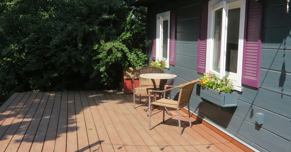 Deck de madera con mesa redonda y sillas de mimbre junto a una casa de fachada gris con ventanas de postigos