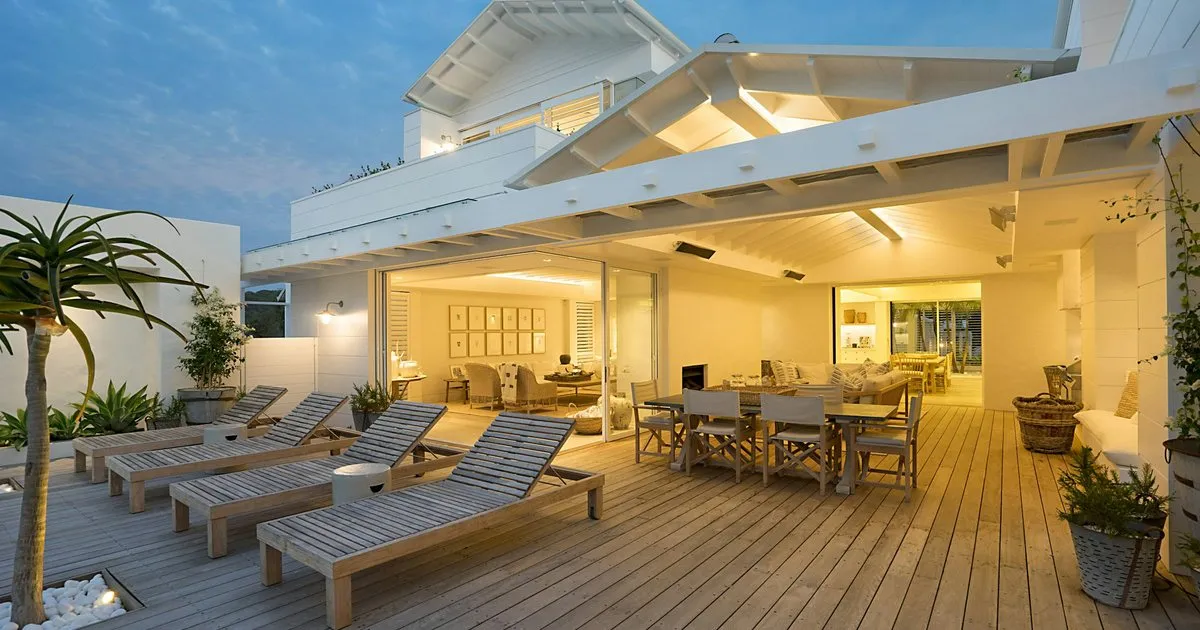 Terraza amplia con deck de madera, asoleadoras y comedor exterior frente a una casa iluminada al atardecer