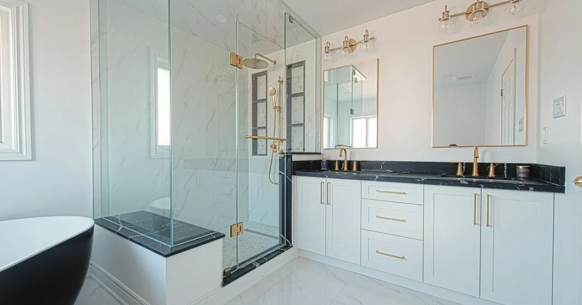 Baño blanco con ducha esquinera cerrada en vidrio templado transparente con herrajes dorados, junto a un mueble de lavamanos blanco con mesón oscuro y dos espejos