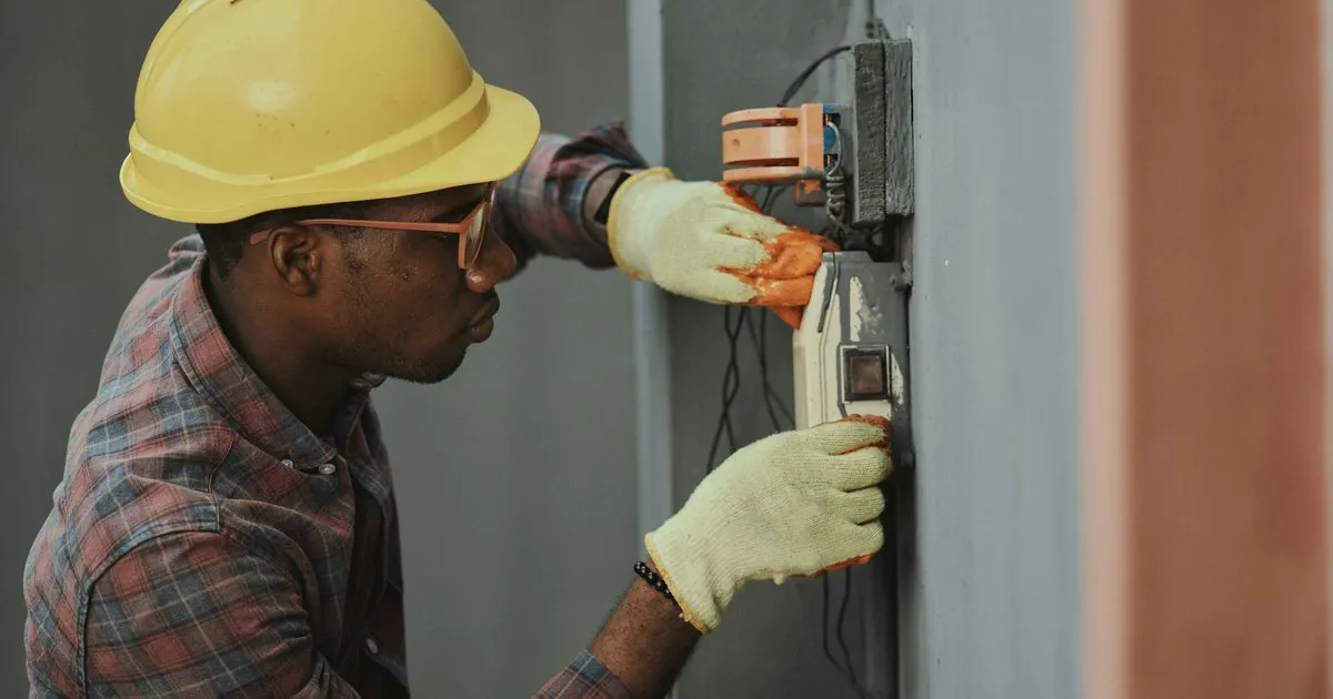 Electricista con casco amarillo y guantes instalando un interruptor en la caja eléctrica de un muro