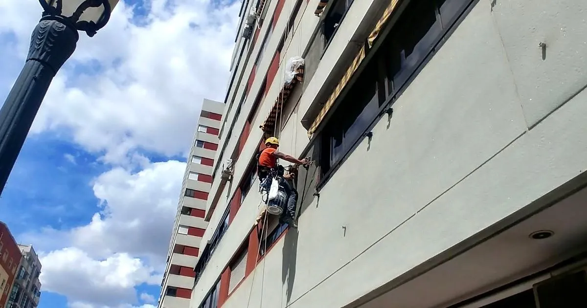 Operario con casco suspendido con cuerdas trabajando sobre la fachada de un edificio de varios pisos