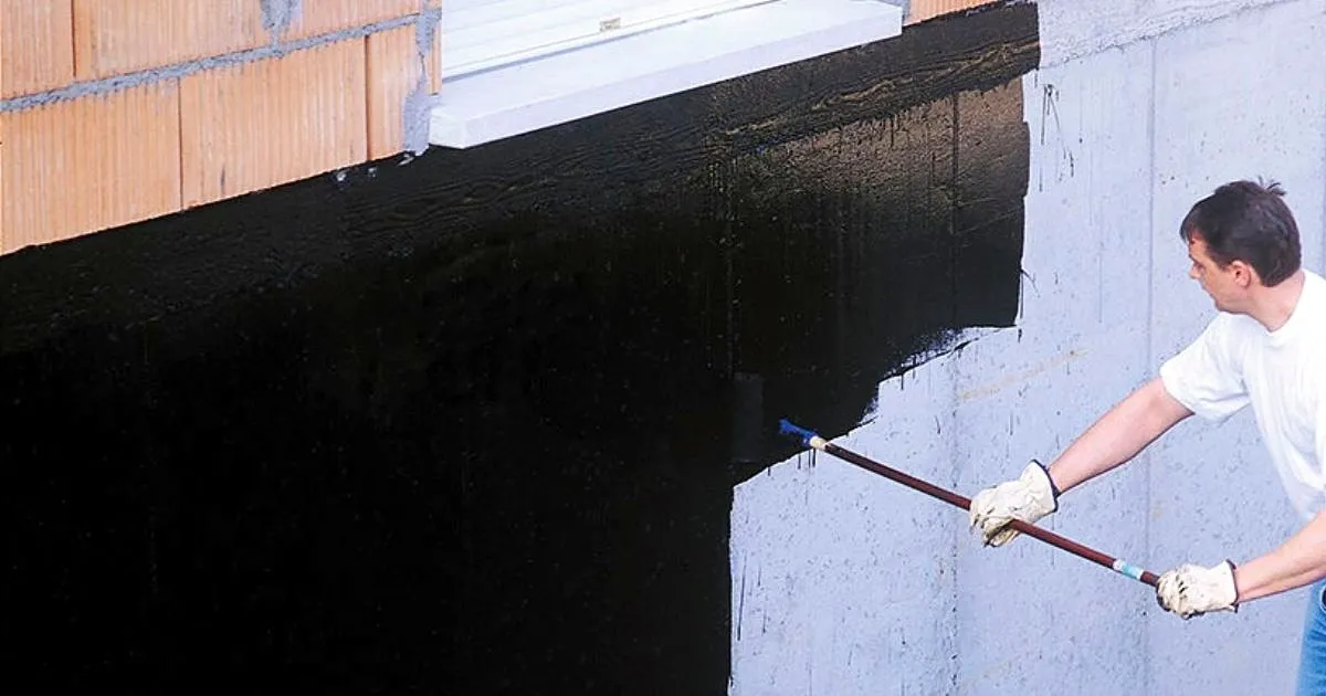 Trabajador con guantes aplicando con brocha de mango largo un impermeabilizante negro sobre un muro exterior de concreto