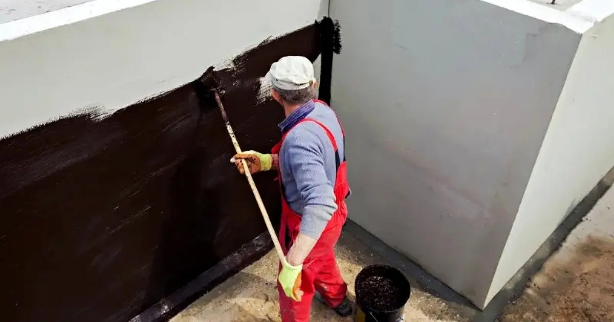 Operario con overol rojo aplicando impermeabilizante negro con rodillo de mango largo sobre la base de un muro