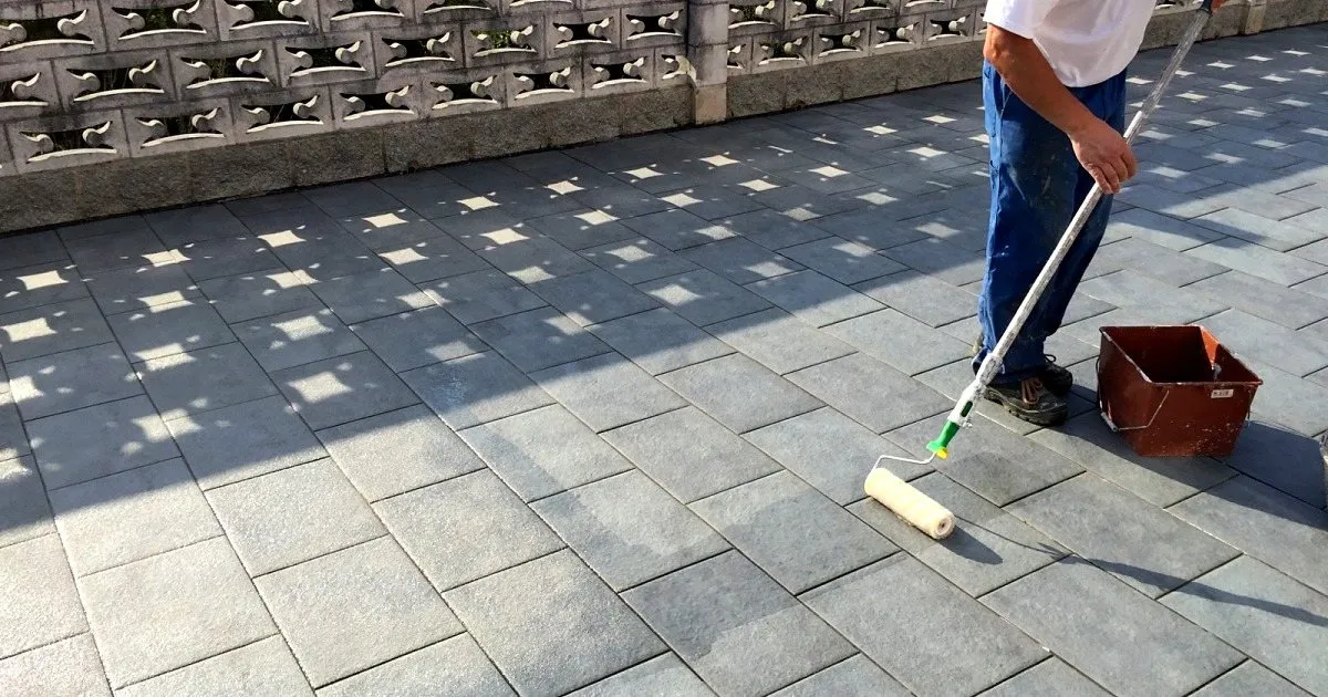 Operario aplicando un producto con rodillo de mango largo sobre una terraza de adoquín gris junto a un muro calado