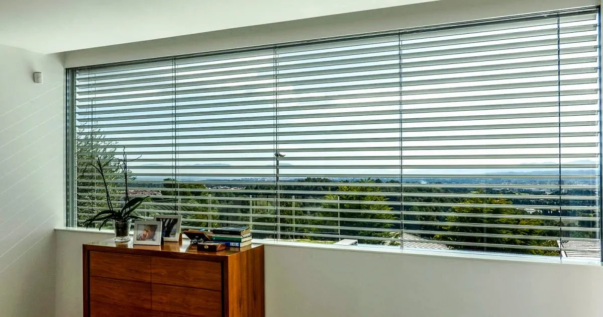 Ventanal panorámico con persianas horizontales claras entreabiertas sobre una cómoda de madera y vista a zona verde