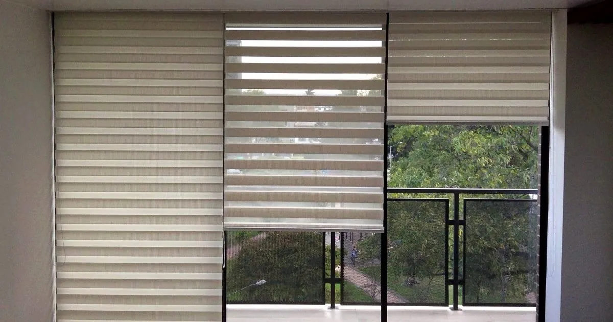 Cortinas de franjas horizontales en tono beige sobre un ventanal, con el paño central recogido y vista a un balcón con árboles