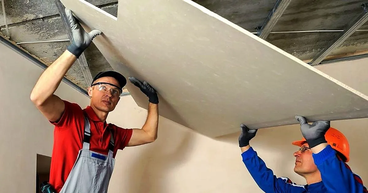Dos instaladores de drywall con casco levantando una placa de yeso hacia la estructura del techo