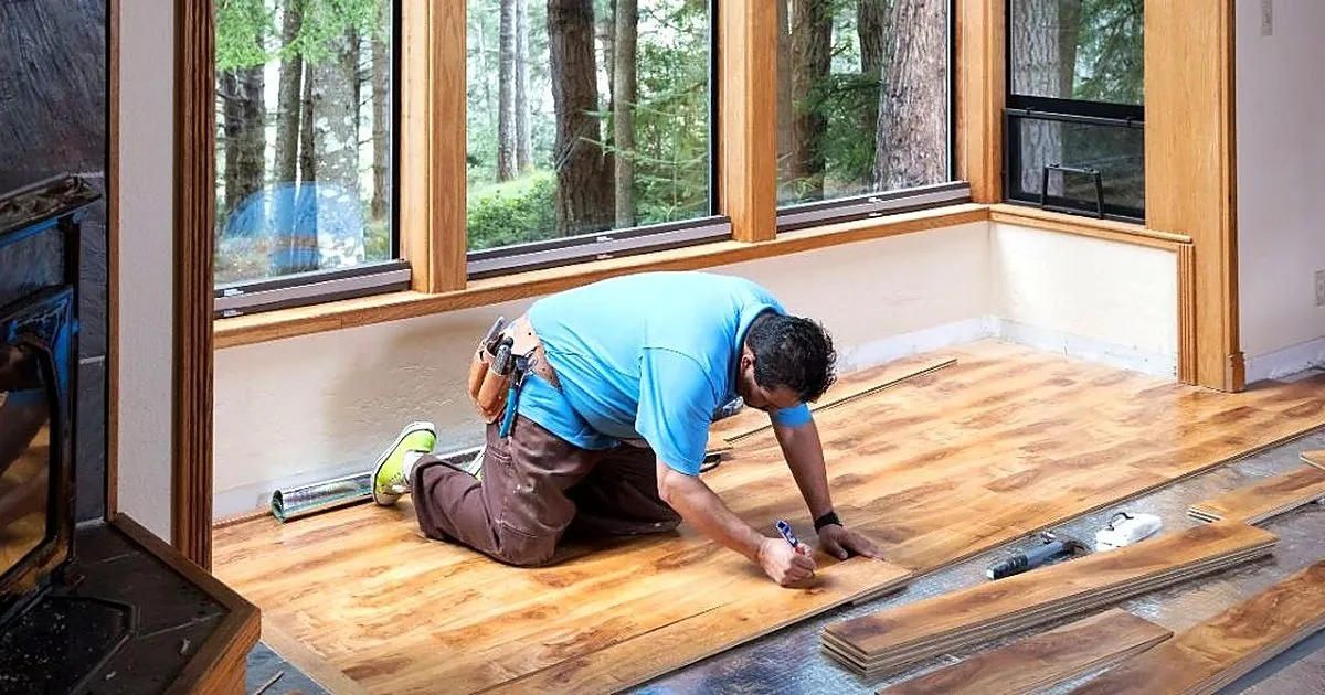 Instalador de camiseta azul fijando tablas de madera de un piso junto a ventanales con vista a un bosque