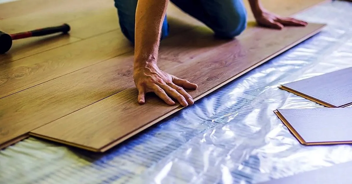 Instalador de rodillas ensamblando tablones de piso laminado tipo madera sobre un manto plateado, con mazo de caucho al lado