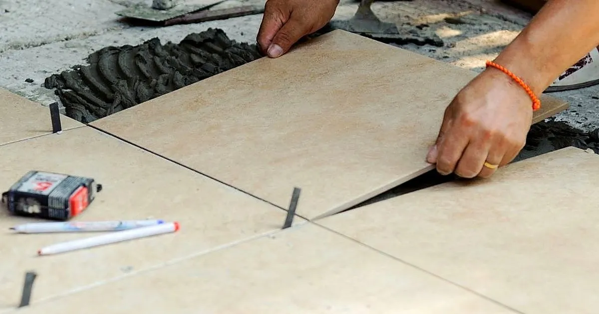 Manos de un instalador asentando una losa de piedra beige sobre mortero peinado, con crucetas en las juntas