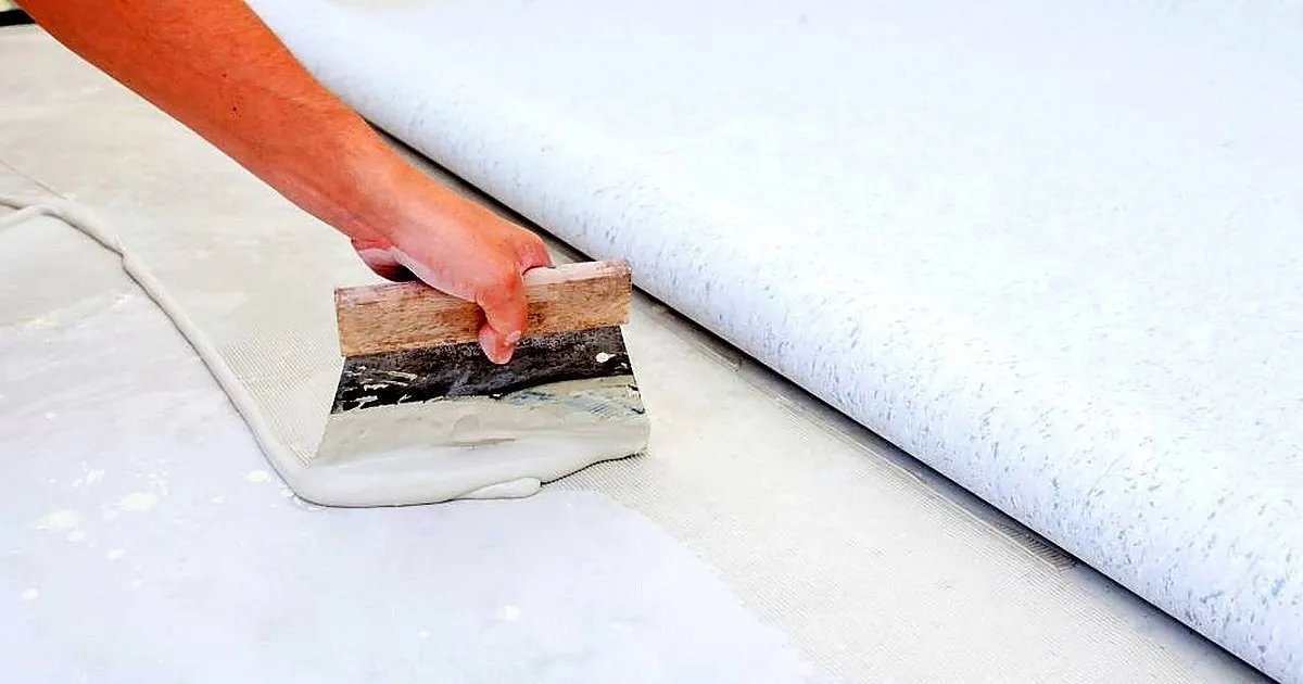 Mano extendiendo adhesivo con una llana sobre la base lisa junto a un rollo de piso PVC claro