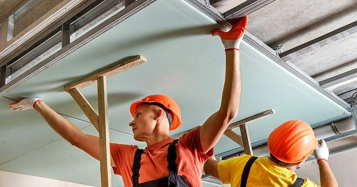 Dos obreros con cascos naranjas y guantes sostienen una placa verde contra perfiles metálicos del techo, apoyada en puntales de madera