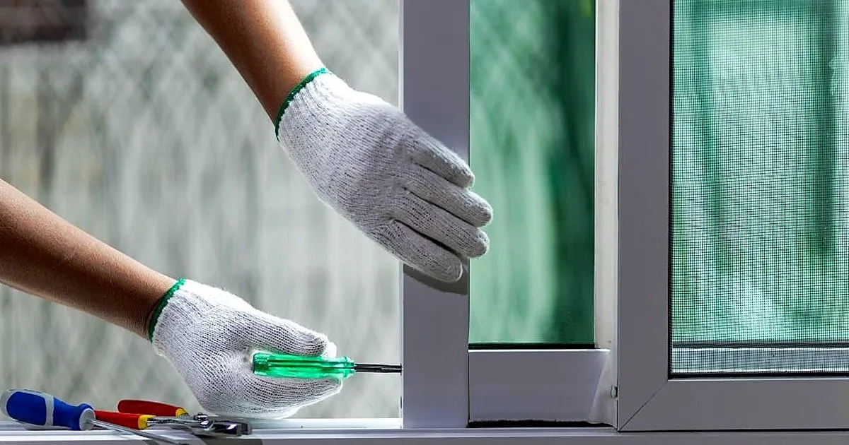 Manos con guantes de trabajo ajustando con destornillador el marco de una ventana corrediza blanca con anjeo