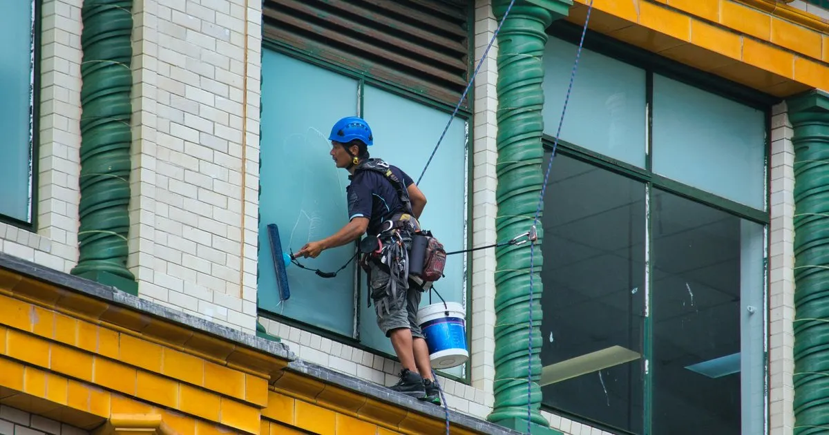Operario con casco y arnés, suspendido con cuerdas, limpiando los vidrios de una fachada de colores con un balde colgado