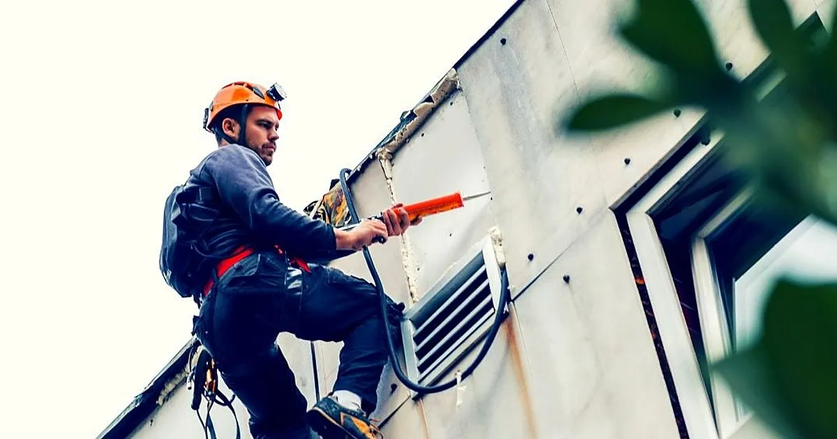 Técnico con casco naranja y arnés, suspendido en la fachada de un edificio, aplicando sellante con pistola junto a una rejilla de ventilación