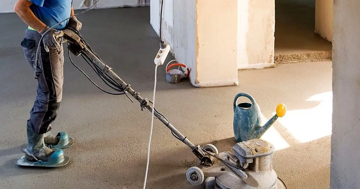 Operario con botas de trabajo pasando una máquina rotativa sobre un piso de cemento gris en un espacio en obra