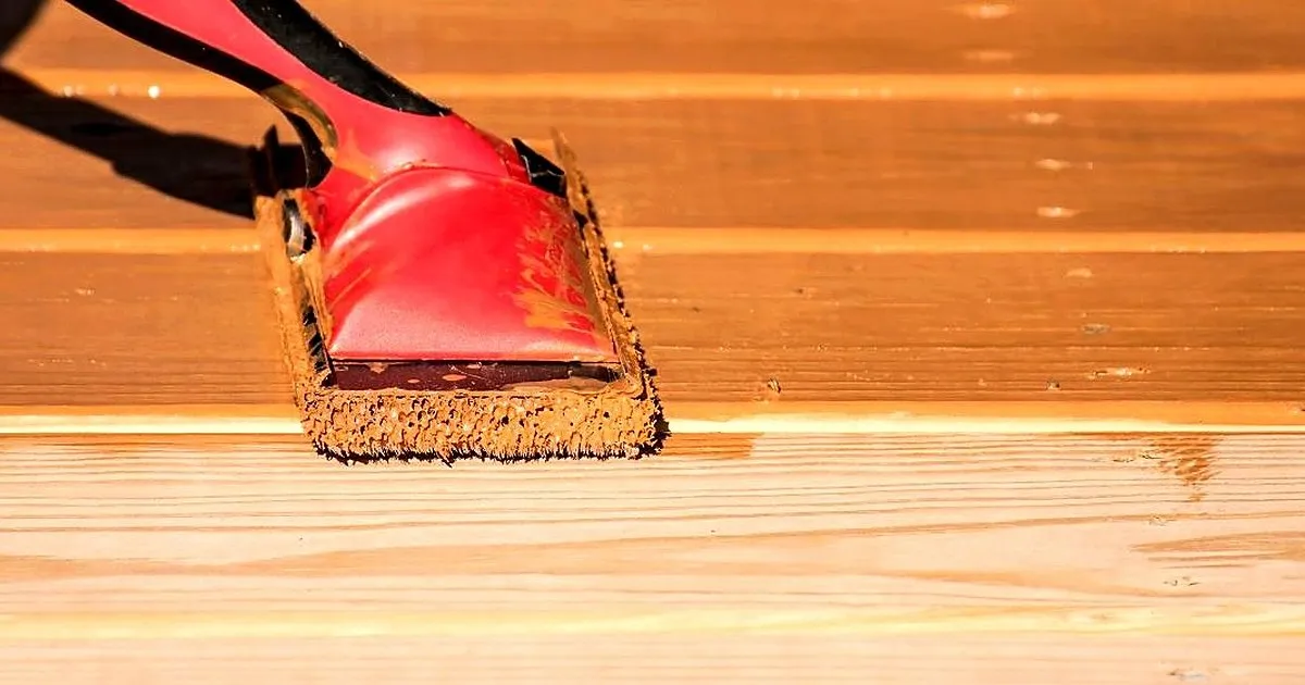 Aplicador de almohadilla con mango rojo extendiendo tinte protector sobre las tablas de un deck de madera