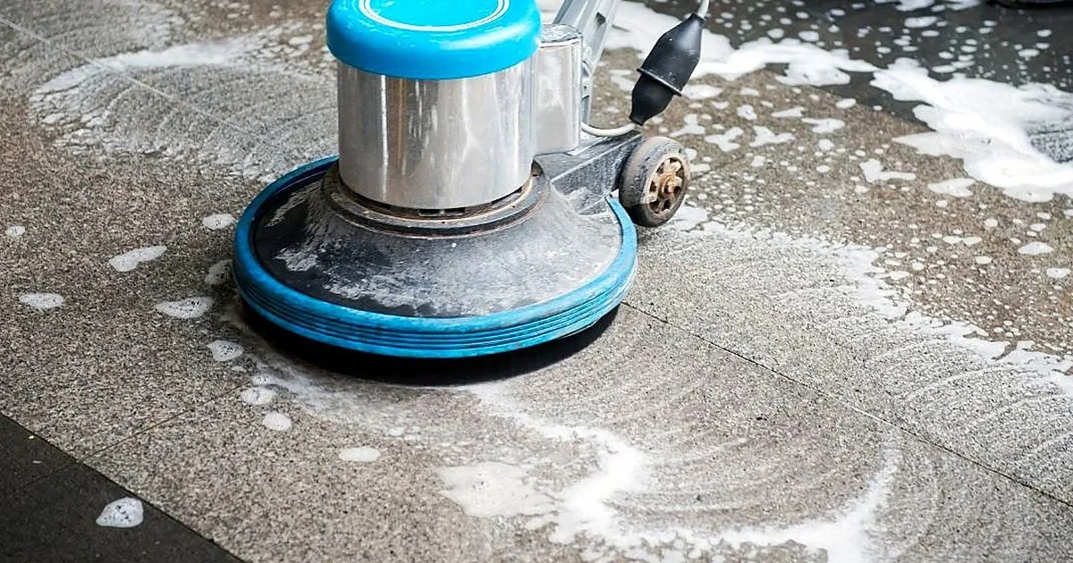 Máquina rotativa con disco azul lavando con espuma un piso de granito gris moteado