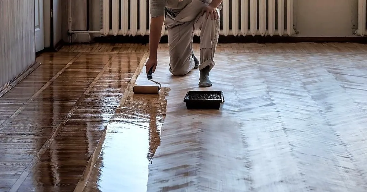 Operario arrodillado aplicando laca con rodillo sobre un piso de parquet en espiga recién pulido