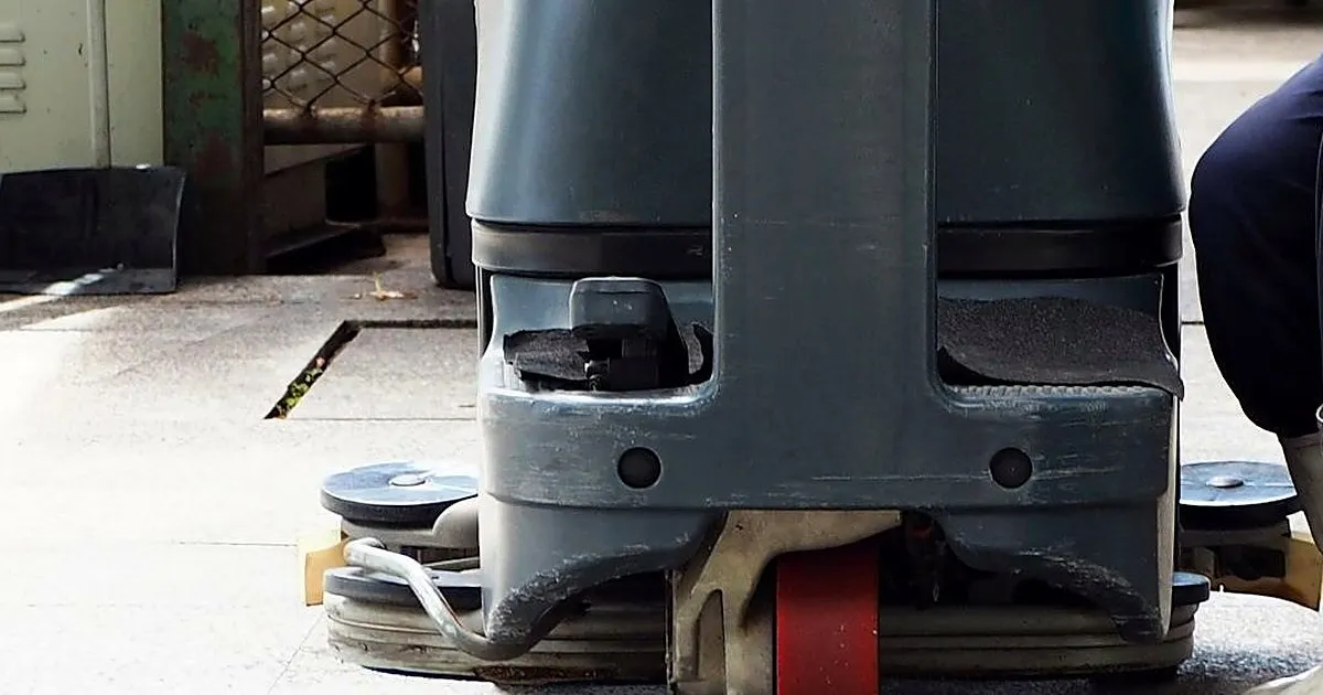 Máquina fregadora industrial de discos limpiando un piso de piedra en un patio, junto a un operario con botas de caucho