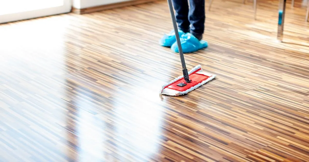 Persona con polainas azules pasando un trapero plano rojo sobre un piso vinílico brillante tipo madera