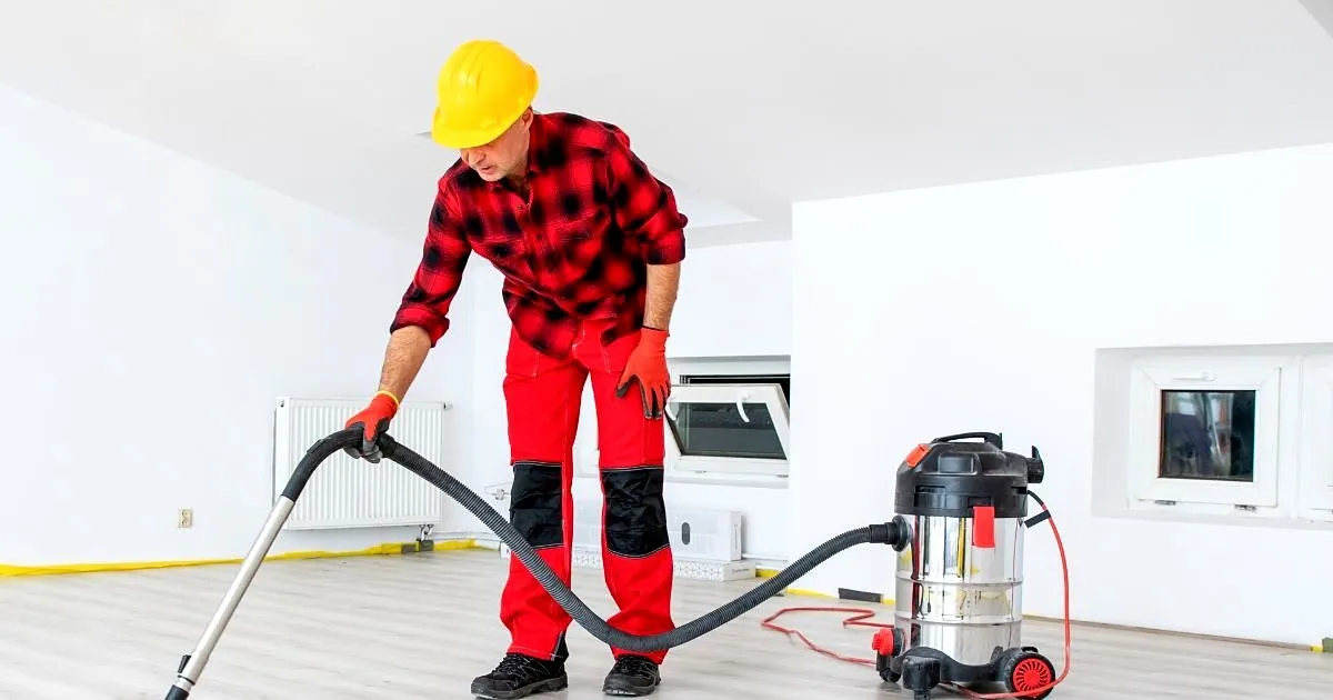Operario con casco amarillo y overol rojo aspirando un piso claro tipo madera en un apartamento blanco