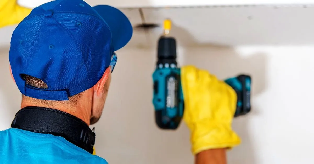 Técnico con gorra azul y guantes amarillos atornillando una lámina de techo en drywall con taladro inalámbrico