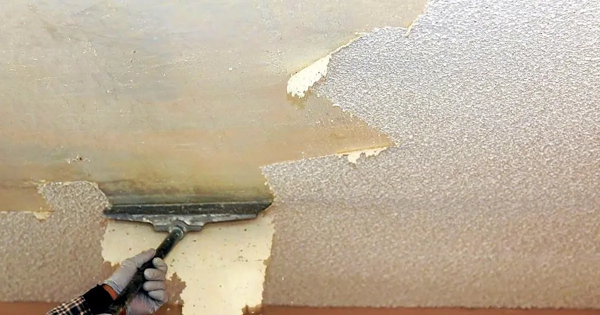 Operario con guantes raspando con espátula ancha la pintura texturizada deteriorada de un techo interior