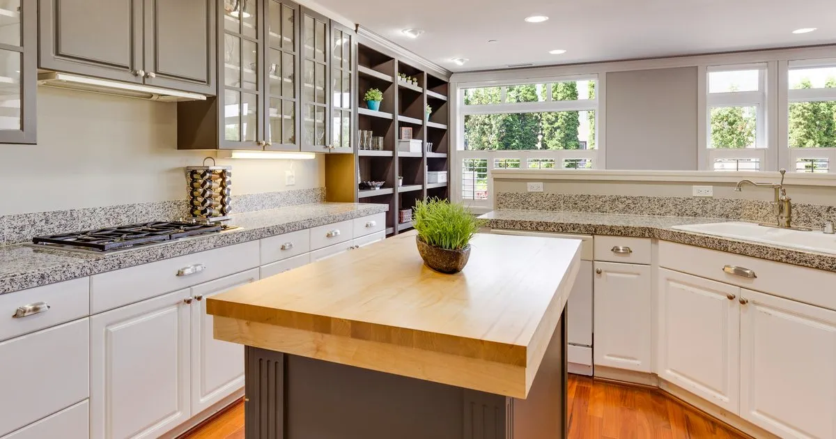 Cocina con mesones de granito gris moteado en L, gabinetes blancos e isla central con cubierta de madera