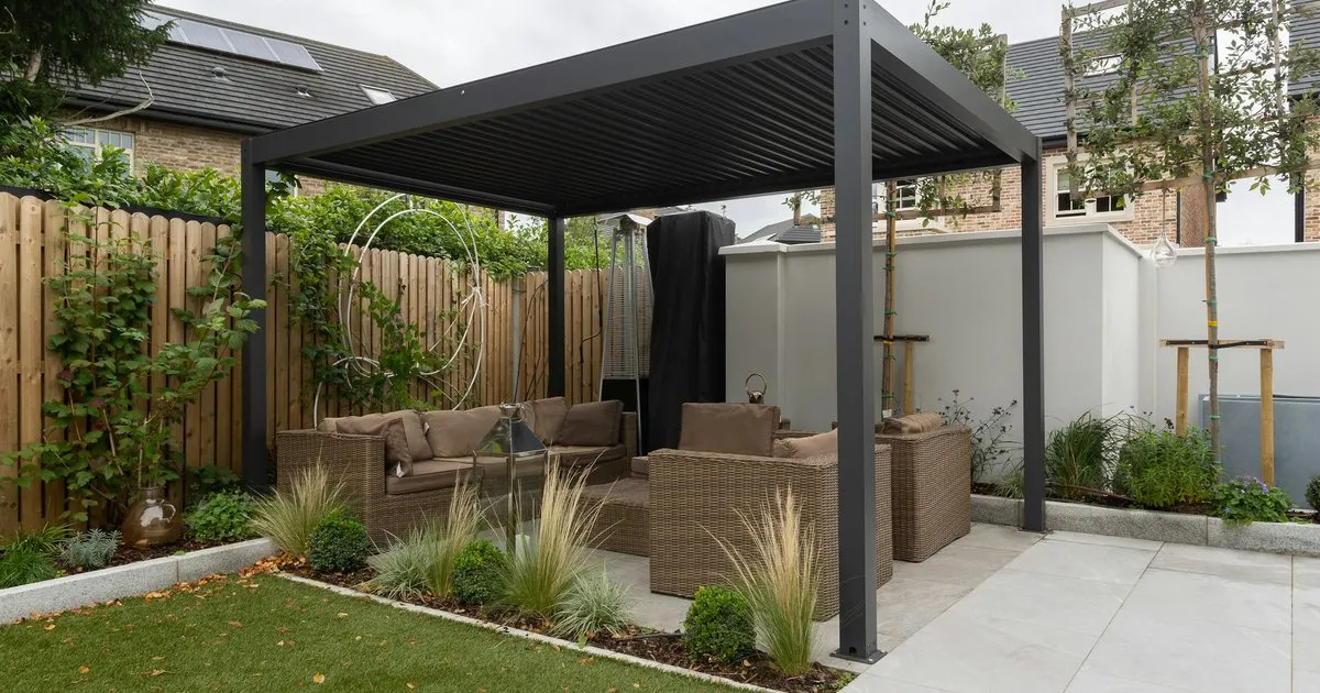 Pérgola metálica negra de cubierta plana sobre una terraza con sala exterior, junto a un jardín con cerca de madera