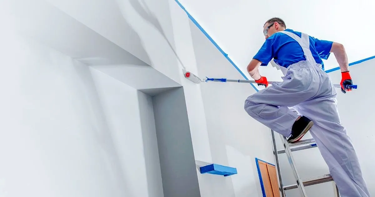 Pintor sobre una escalera pintando con rodillo de extensión un muro alto delimitado con cinta de enmascarar azul