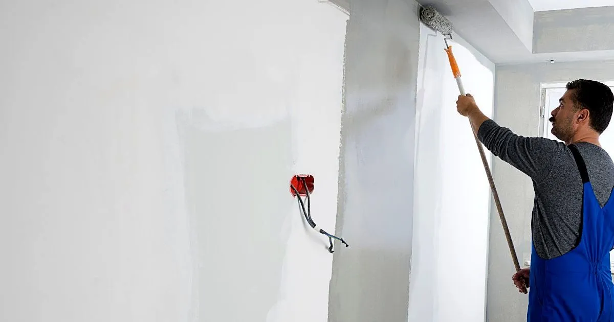 Pintor con overol azul aplicando pintura blanca en un muro con rodillo de extensión