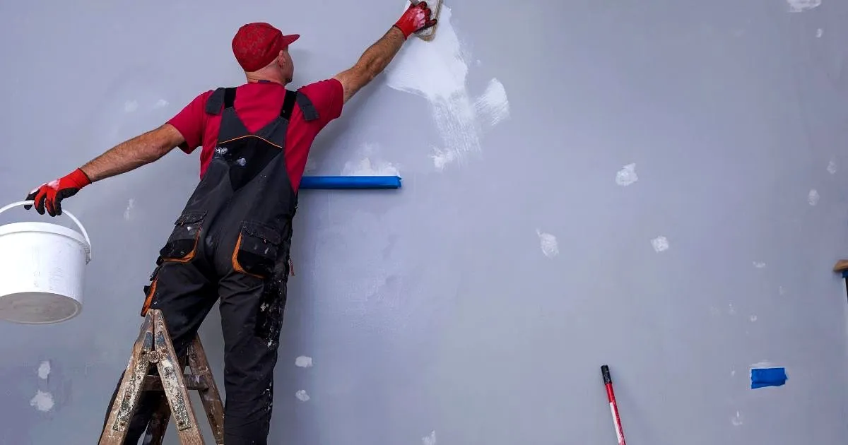 Operario sobre una escalera resanando una pared gris de un local, con balde blanco en la mano