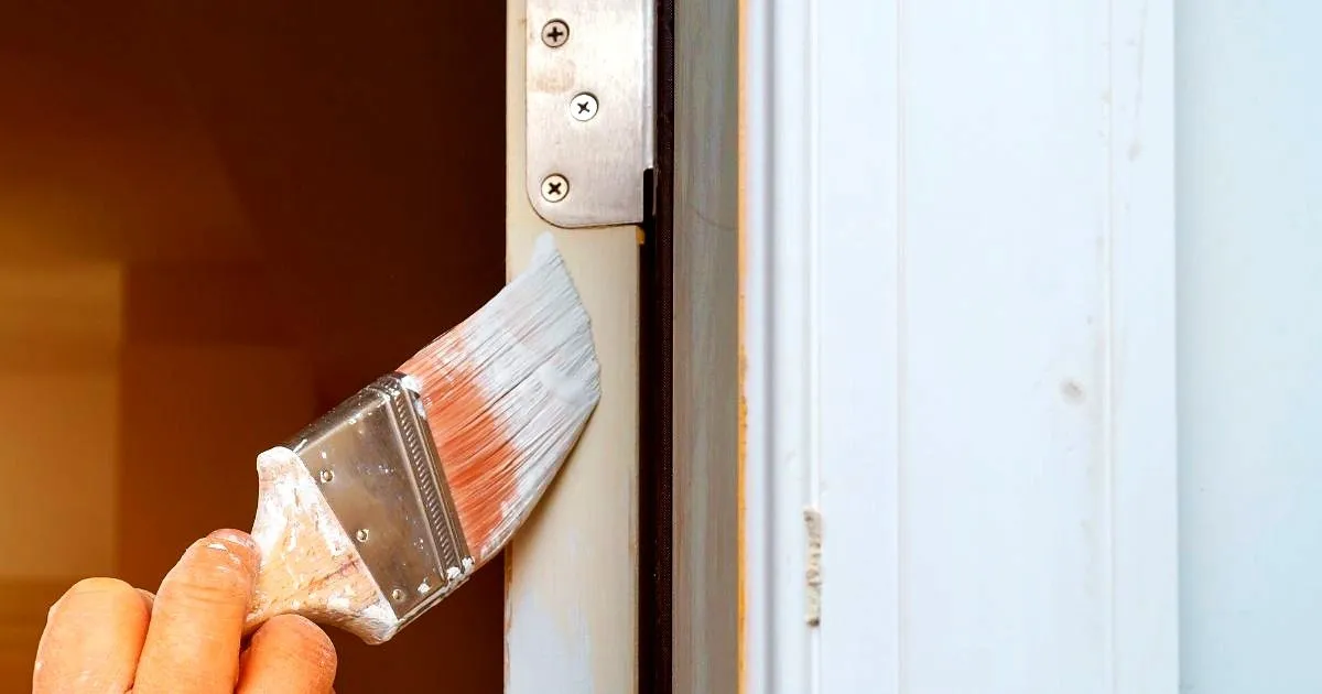 Brocha aplicando pintura blanca sobre el marco de una puerta, junto a la bisagra
