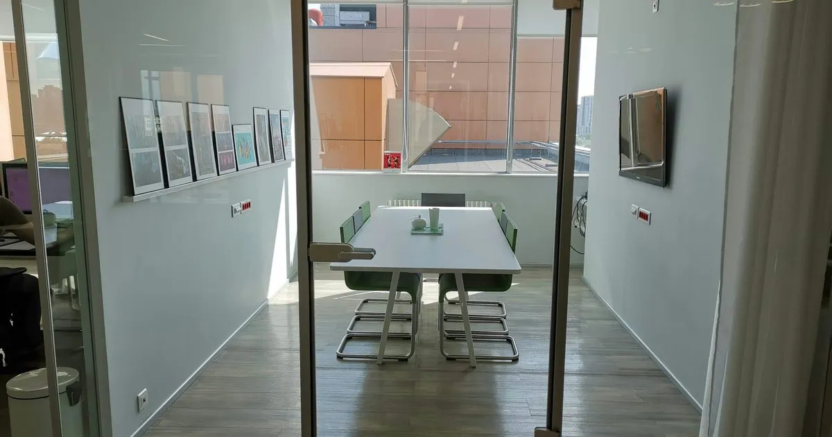 Puerta de vidrio abierta hacia una sala de reuniones con mesa blanca y sillas, entre muros con cuadros enmarcados