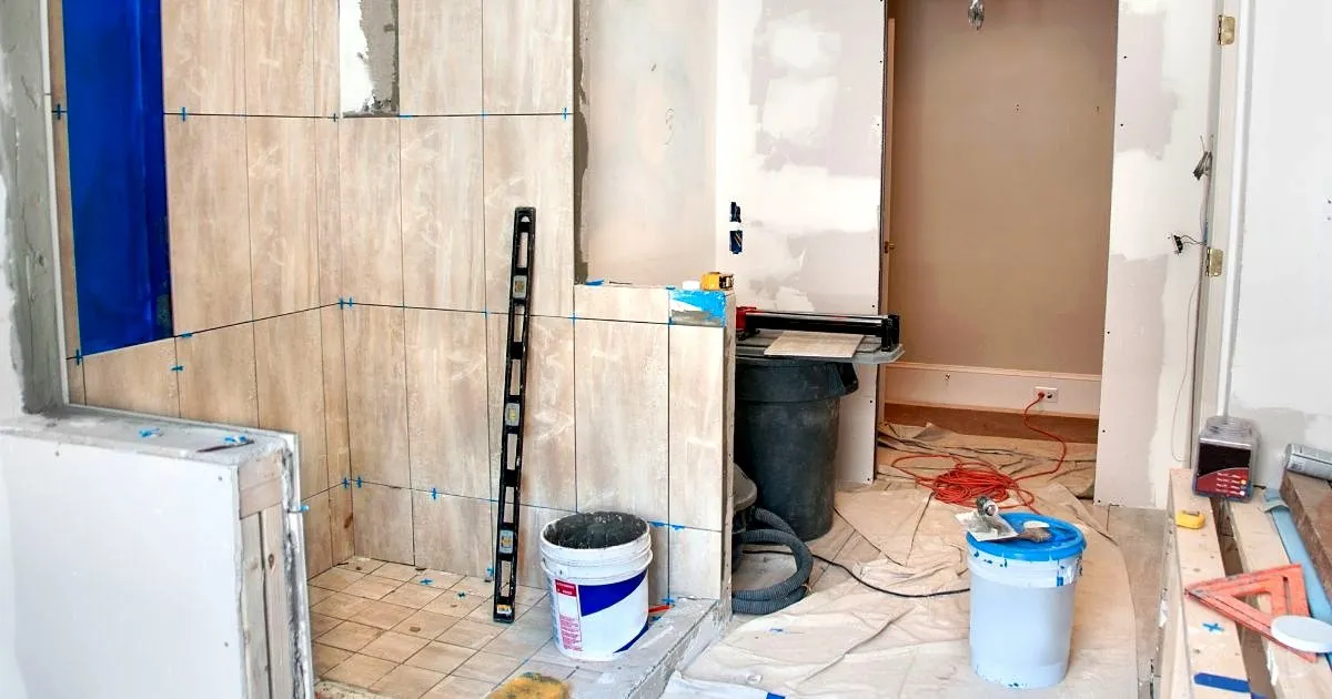 Ducha en remodelación con enchape beige de gran formato a medio instalar, crucetas azules, nivel apoyado en el muro y baldes de pegante