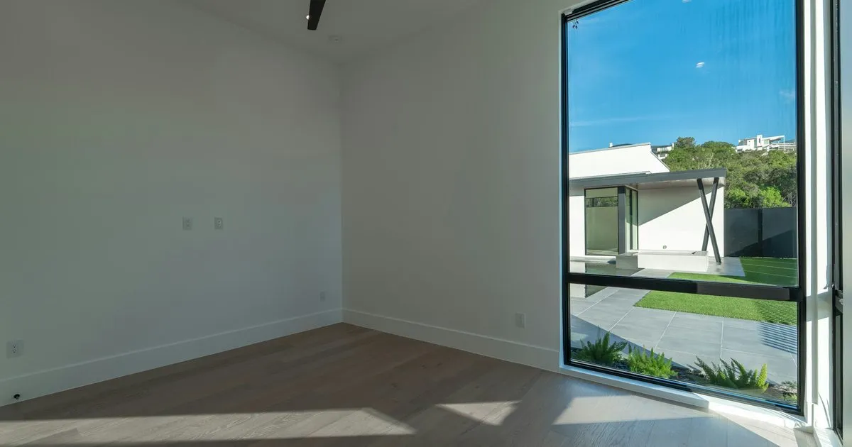 Habitación vacía de muros blancos con una ventana esquinera de aluminio negro y vista a un patio